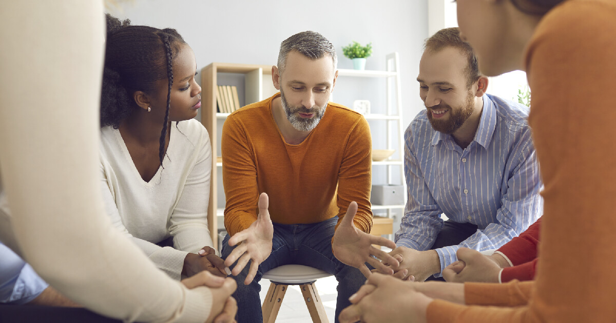 A diverse group of people sits in a supportive circle, listening intently as a man in an orange sweater speaks during a group therapy session.