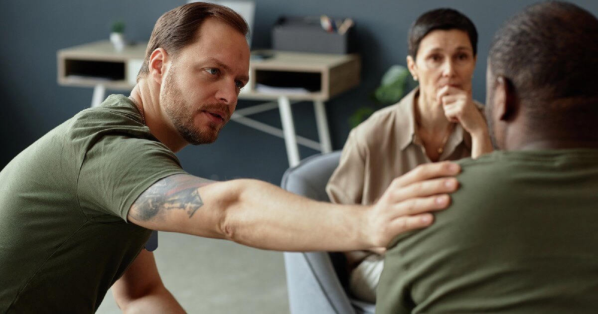 A man offers a reassuring hand on another man's shoulder during a trauma-focused group therapy session, while a woman observes thoughtfully.
